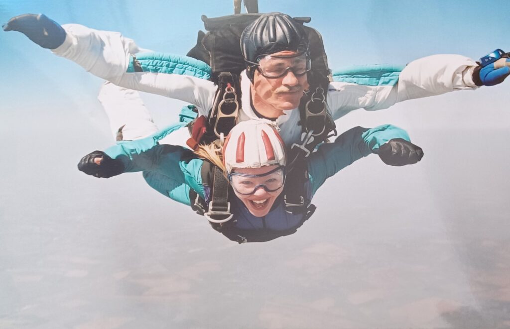A photograph of a tandem skydive. Toni is attached to the instructor - they both have their arms out and are smiling.