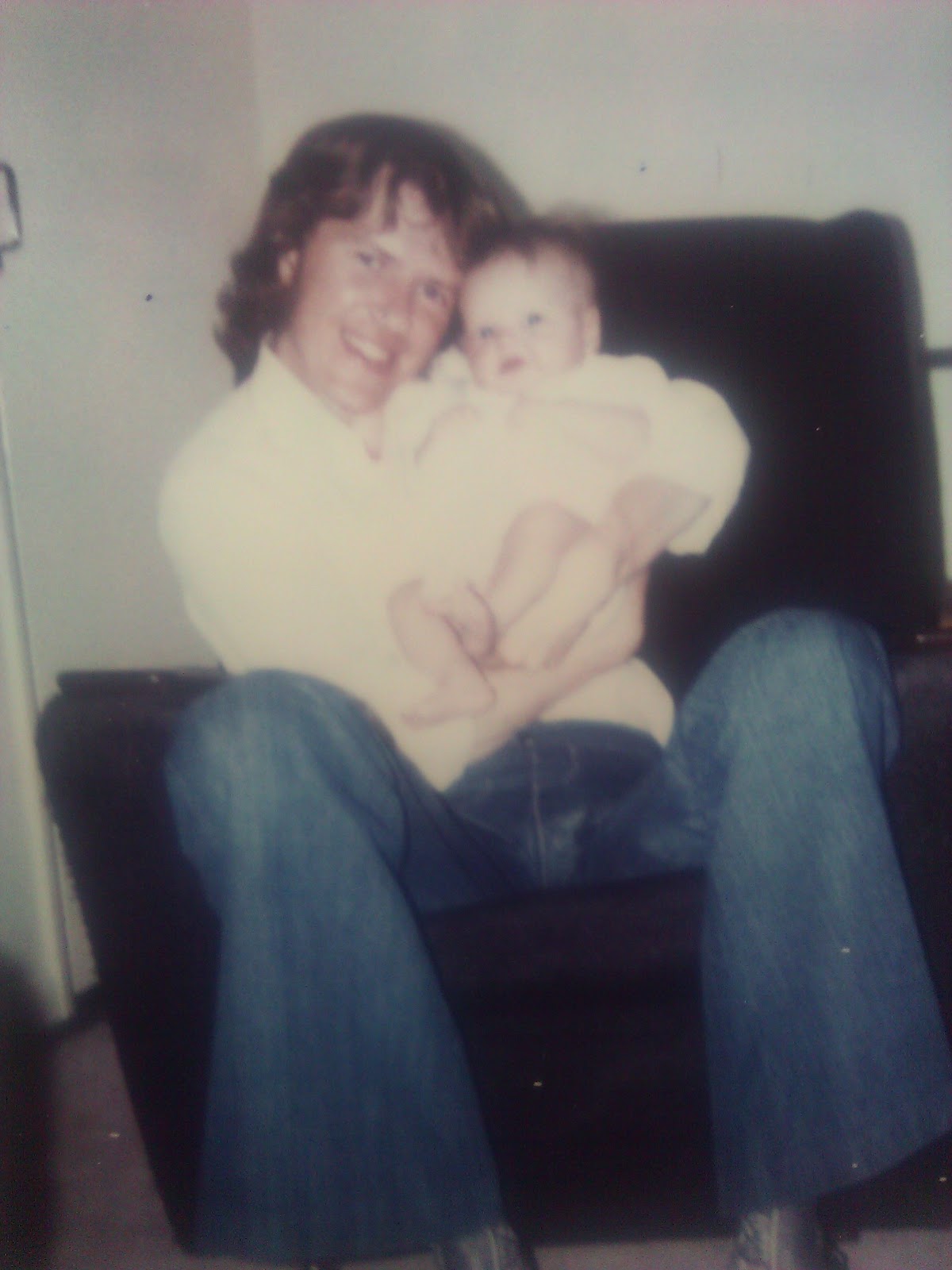 a picture of Toni's dad holding her when she was around 6 months old. he is sat in a chair and wearing blue denim flared jeans and a white shirt