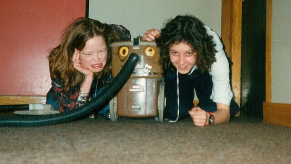 Two teenage girls crouched either side of a vacuum cleaner. One is smiling, the other one is pulling a face as it she is sad.