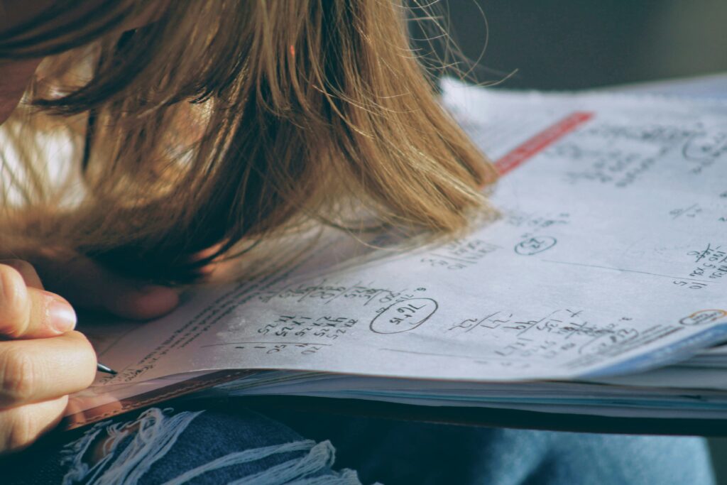 A child's head is bowed over an exercise book with mathematical calculations written in it. you can only see the child's hair and the top of their right hand which is holding a pencil